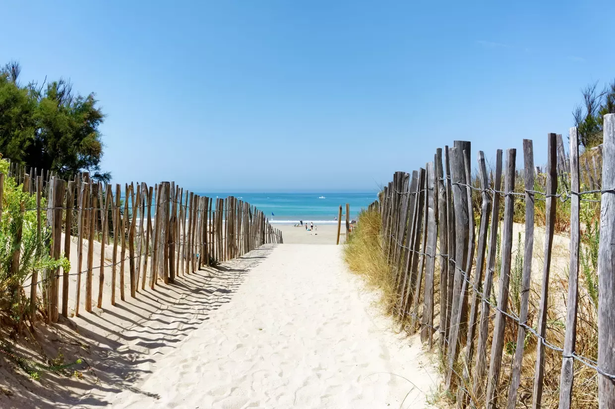 Un chemin de sable entre les ganivelles mène à la plage, l’océan turquoise au bout